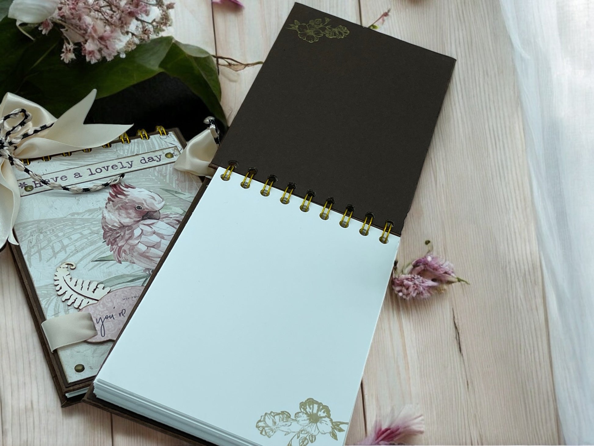 Flat lay of A6 cockatoo notebook with golden spiral binding, designer paper cover and ribbon bow—handmade journal for everyday notes and sketches.